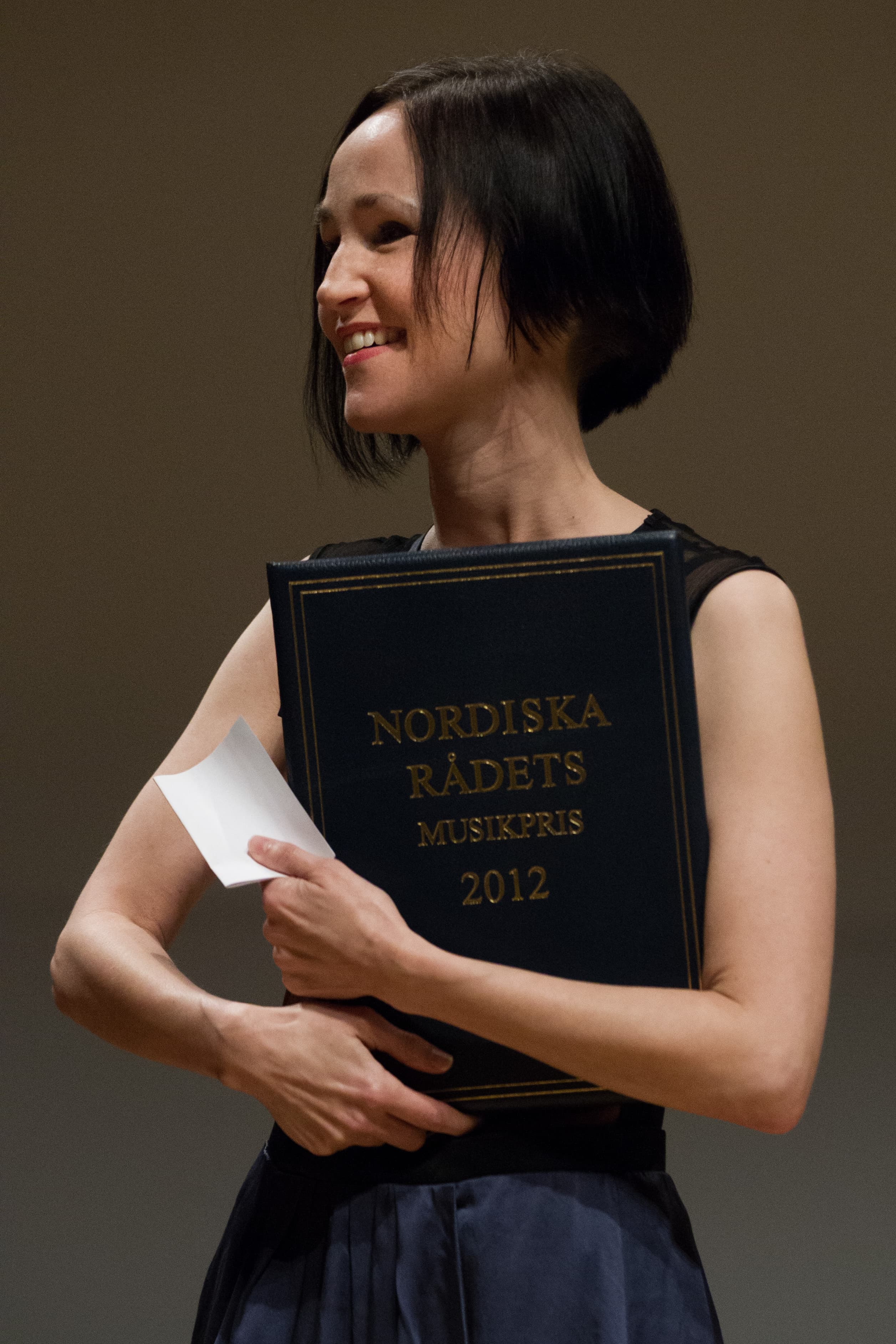 A person is smiling while holding a book titled "Nordiska Rådets Musikpris 2012."