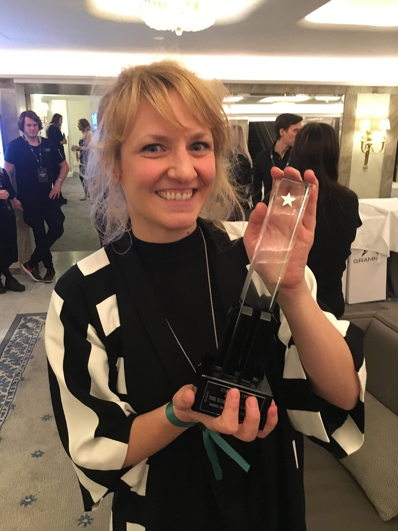 A smiling person is holding a trophy with a star on it in a well-lit room with several others in the background.