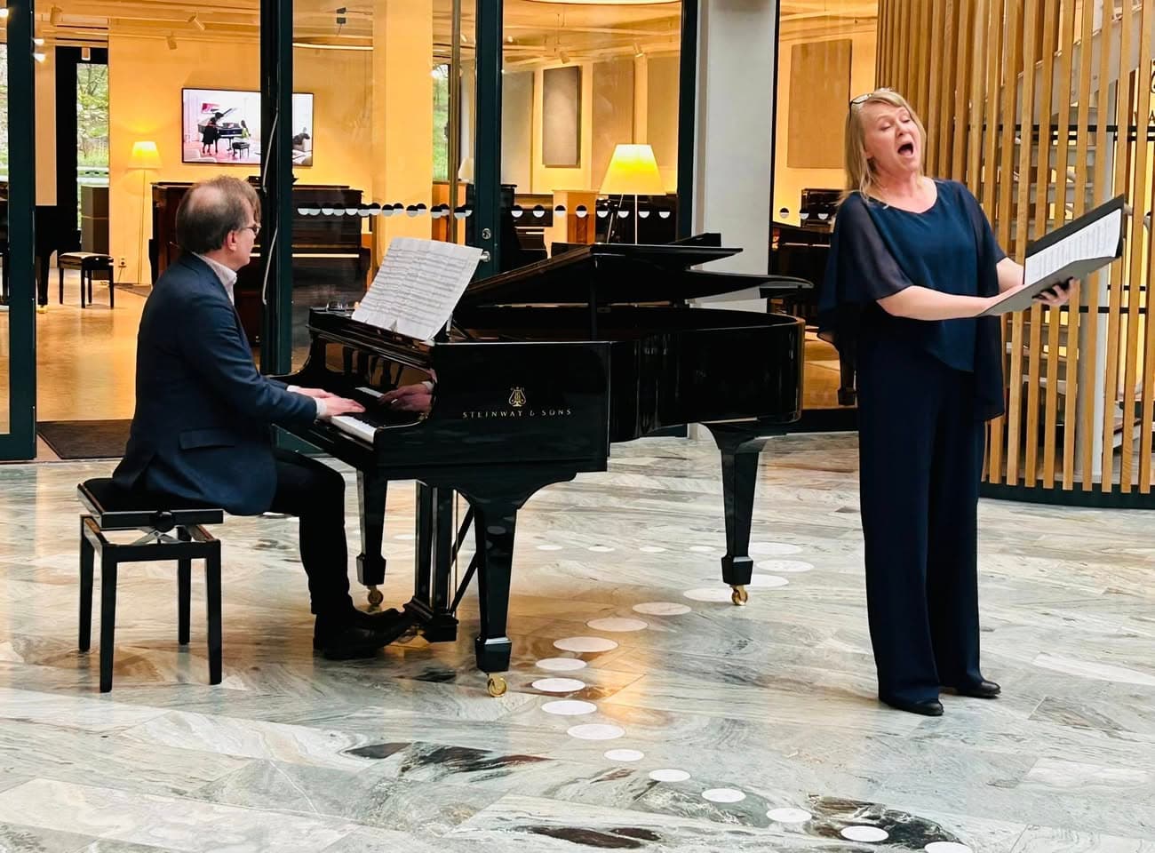 A pianist is playing a grand piano while a woman stands nearby singing with sheet music in hand.